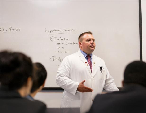 Physician in a white coat giving a lecture to medical students, standing in front of a whiteboard with handwritten notes.
